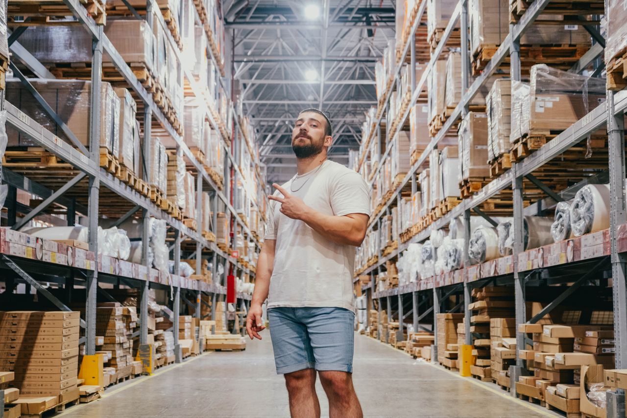 services-01 Young man posing in a warehouse aisle surrounded by racks and packages inside a spacious store.
