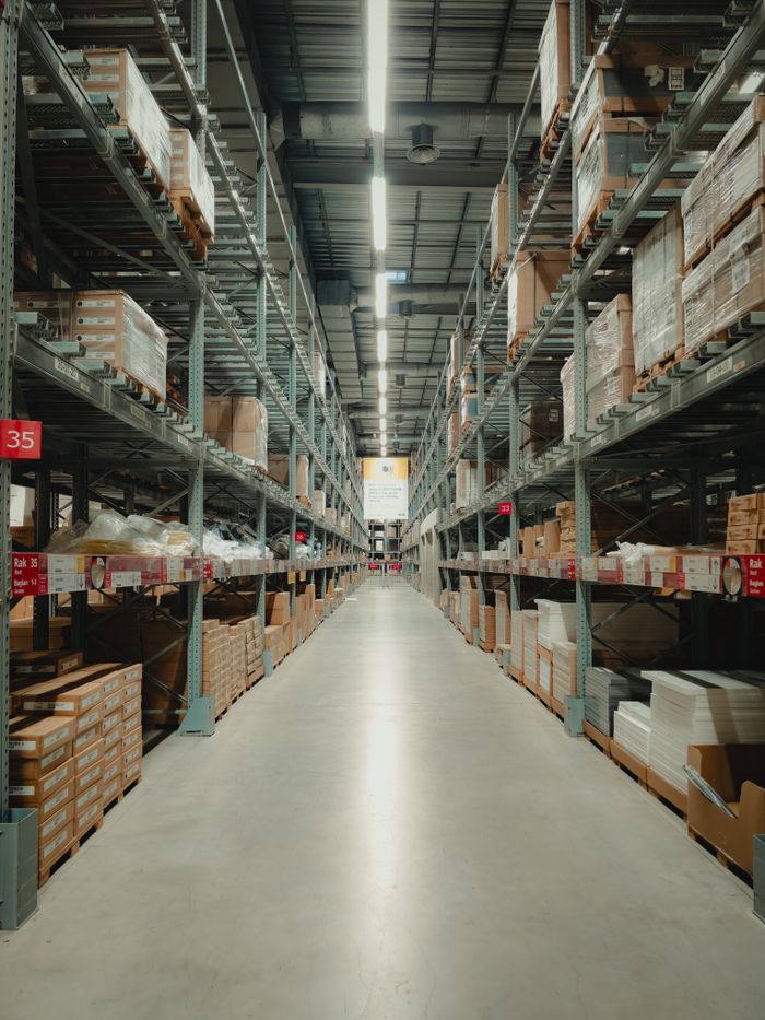 about-01 Spacious warehouse aisle with shelves filled with neatly stacked products and pallets.